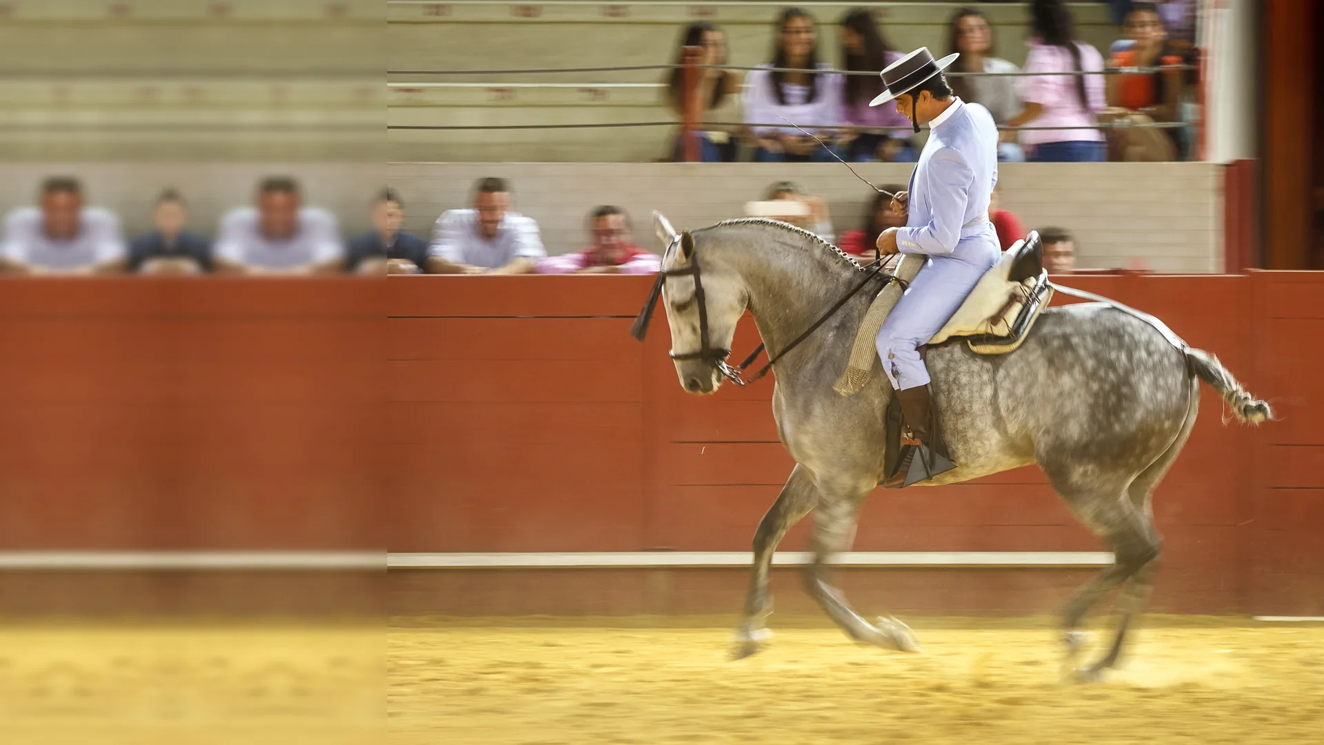 A rider in a pale blue jacket and flat-brimmed hat on a dapple-grey horse moving across the sand of an arena, with spectators behind a red wall