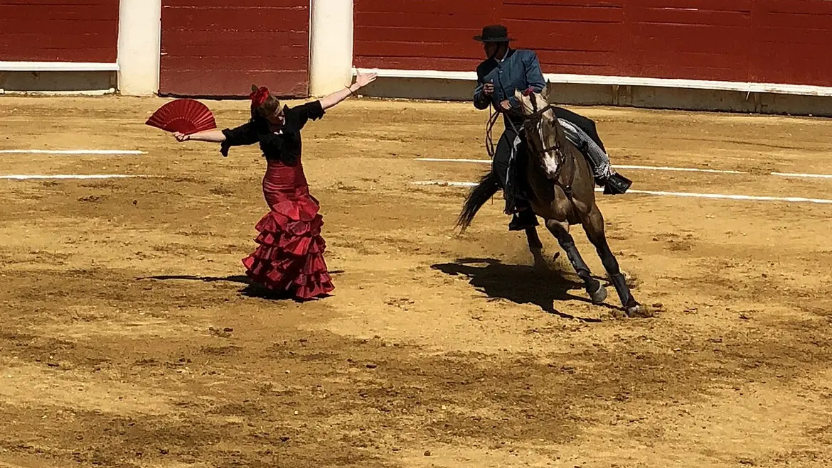 A flamenco dancer in a red ruffled skirt holds out a red fan on the sand of an arena while a rider on a dark horse circles beside her
