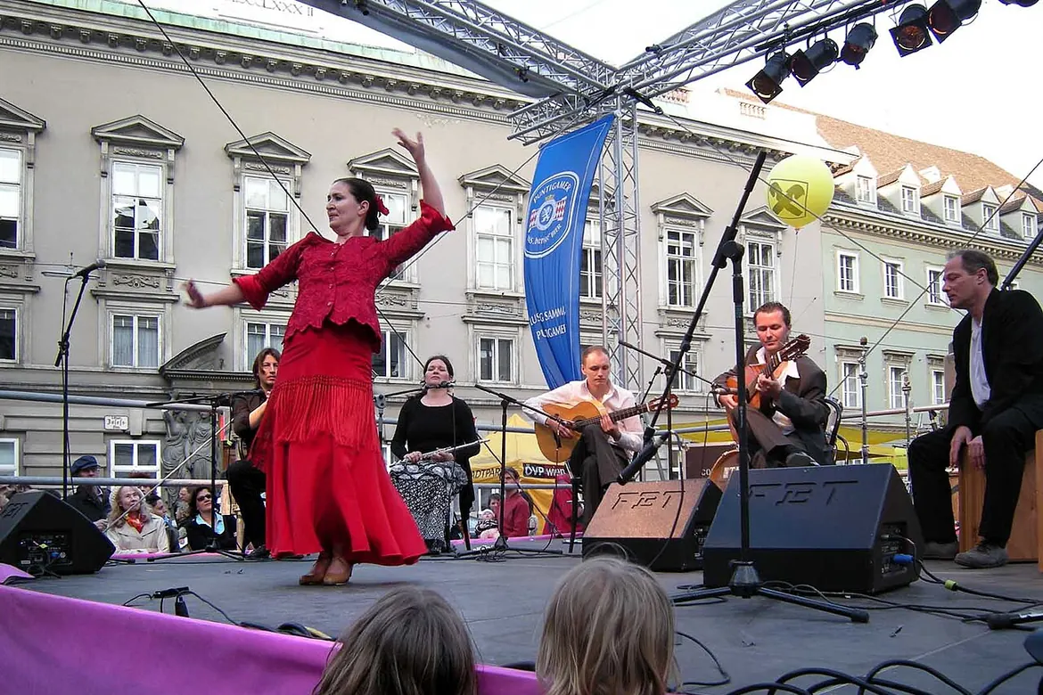 On a festival stage a dancer in a red dress strikes a pose, with two guitarists and a singer seated behind her